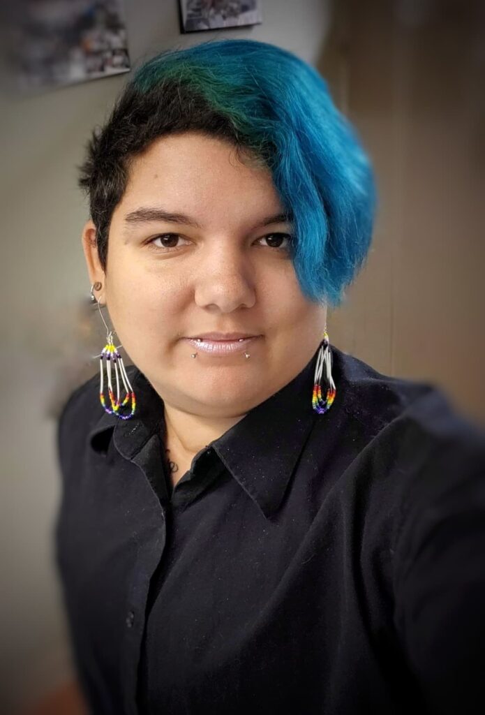 A person with short blue and black hair is looking at the camera. They are wearing a black shirt and chandelier earrings. 